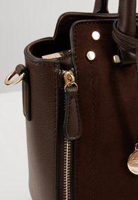 Coin d’un sac à main en cuir marron foncé montrant une fermeture éclair dorée, des coutures, des rivets métalliques, une attache anneau et un charm rond doré de marque.