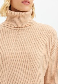 Beżowy sweter typu golf z prążkowaną strukturą, ozdobiony diagonalnymi wzorami i miękką fakturą. Zaprojektowany dla ciepła i komfortu z dopasowanym kołnierzem.