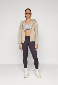 Žena v béžovej zipsovej mikine, vzorovanom športovom podprsenke, čiernych legínach, bielych ponožkách, béžových teniskách a slnečných okuliaroch stojí na pozadí s jednoduchým vzorom.