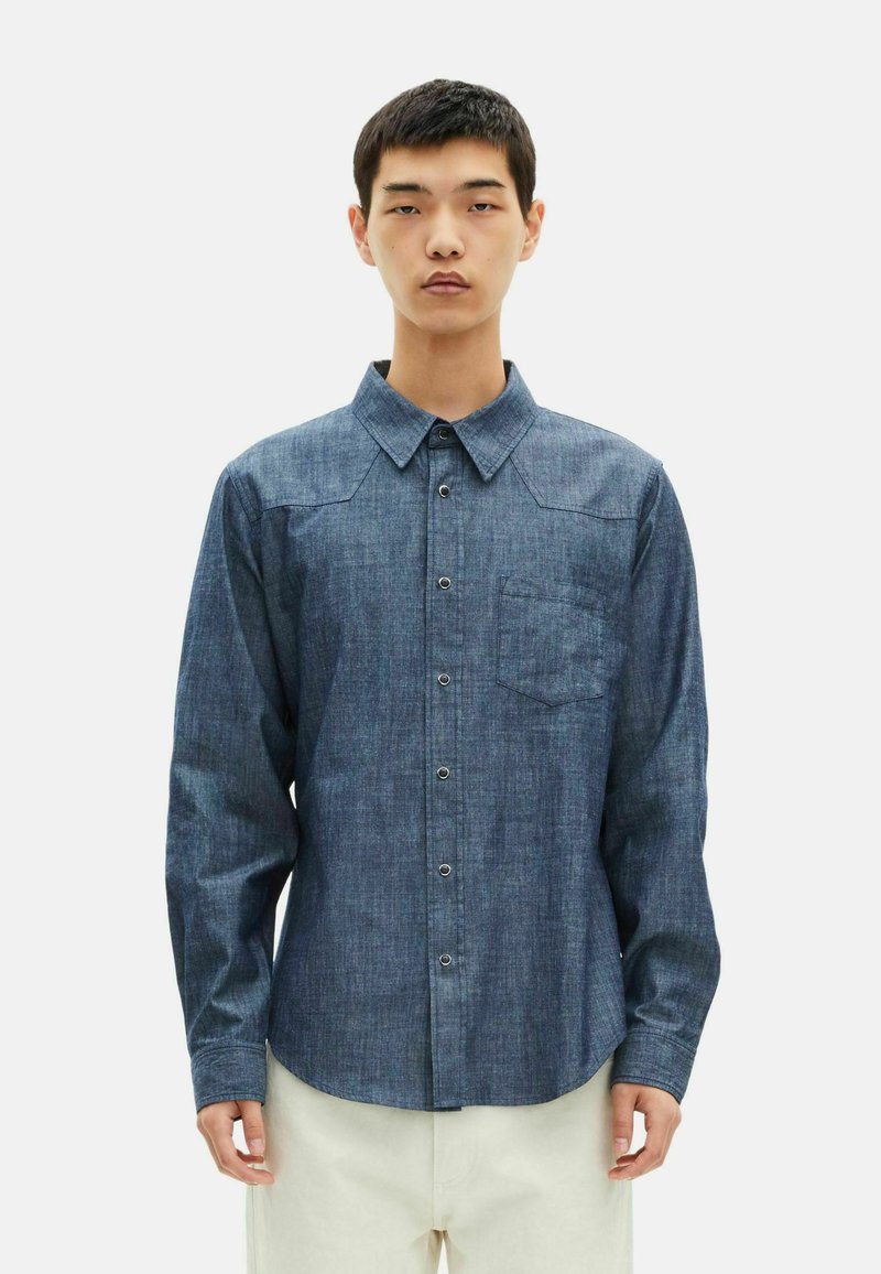 Camisa azul escura de botões em tecido chambray, com mangas longas, colarinho, um bolso no peito e botões em contraste.