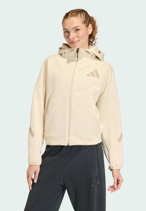Nuori nainen, jolla on beige vetoketjullinen huppari ja mustat logolliset housut, seisoo lievä hymy kasvoillaan ja rentona asennossa.
