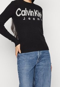 Pull en maille noire avec un logo blanc bien visible "Calvin Klein Jeans" sur le devant, doté de poignets et d'un ourlet côtelés, associé à un jean bleu.