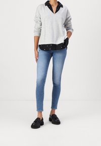 Felpa grigia chiara a V sovrapposta a una maglietta nera con pois bianchi, abbinata a jeans skinny blu e scarpe nere.