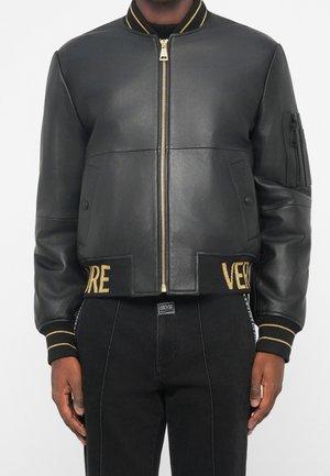 Schwarze Leder-Bomberjacke mit goldenem Reißverschluss, gestreiften Bündchen und "VERSACE" in Gold auf dem Bund bestickt, getragen mit schwarzen Hosen.
