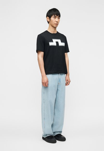 Άνδρας που στέκεται φορώντας μαύρο μπλουζάκι με λευκό γεωμετρικό σχέδιο, ανοιχτό μπλε τζιν παντελόνι με φαρδιά πόδια και μαύρα αθλητικά παπούτσια σε απλό φόντο.