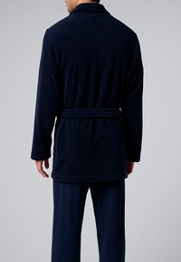 Uomo che indossa una vestaglia in pile blu scuro con cintura legata sul retro, abbinata a pantaloni larghi dello stesso colore, in piedi contro uno sfondo semplice.