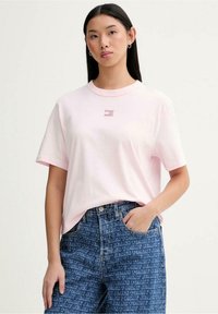 Maglietta rosa in cotone con maniche corte e scollo girocollo, con un piccolo logo sul petto. Abbinata a jeans di denim con motivi testuali.