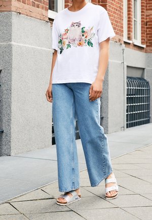 Person iført hvid t-shirt med katte- og blomstermønster, lyseblå vidde bukser og hvide sandaler, der står på en byfortov.