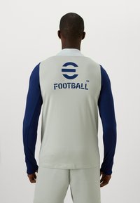 Sportshirt in lichtgrijs met marineblauwe lange mouwen. Kenmerkt zich door het woord "FOOTBALL" in vetgedrukte marineblauwe tekst op de achterkant. Gladde textuur.