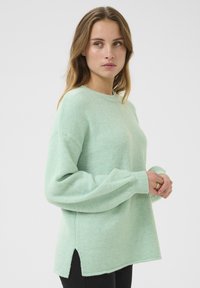 Pull oversized vert clair en matériau doux, avec un col rond et des fentes sur les côtés à l'ourlet pour un détail supplémentaire.