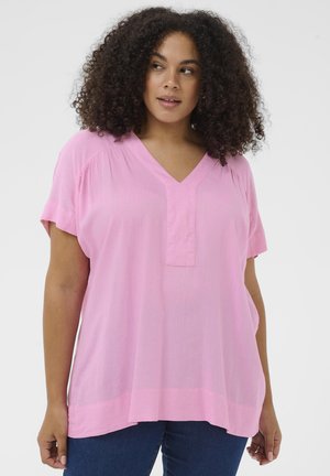 Žena s kučeravými vlasmi, ktorá má na sebe voľné, svetloružové tričko s krátkym rukávom a výstrihom do V a tmavomodré džínsy, pozerajúca sa mierne do strany.