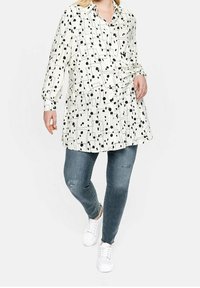 White shirt with black geometric and polka dot pattern, long sleeves, button-up front, paired with distressed blue jeans and white sneakers.