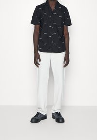 Zwart shirt met korte mouwen en wit patroon, gecombineerd met lichtgrijze broek en zwarte schoenen. Minimalistisch ontwerp, knoopsluiting aan de voorkant, reguliere fit.
