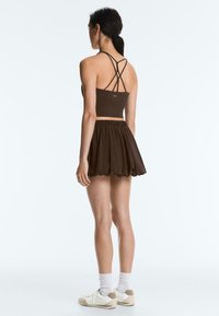 Femme debout, de dos, portant un crop top marron à bretelles, une jupe boule assortie, des chaussettes blanches et des baskets blanches sur un fond uni.