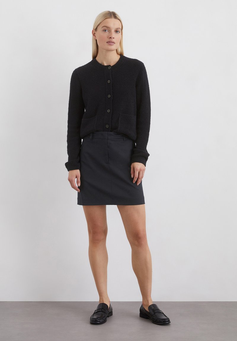 Schwarzer strukturierter Cardigan mit rundem Halsauschnitt, Knopfverschluss und zwei vorderen Taschen. Kombiniert mit einem dunklen Rock und schwarzen Slippers.