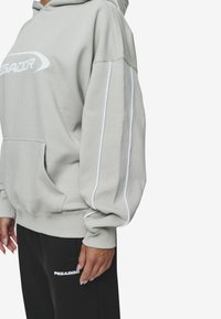Gray hooded sweatshirt with white outline accents on sleeves, kangaroo pocket, and front "PEGADOR" logo. Paired with black pants.