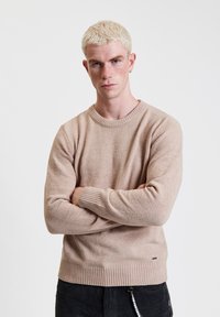 Jasno beżowy sweter z dzianiny z prążkowanymi mankietami i dołem. Prosty, okrągły dekolt. Materiał wydaje się miękki i teksturowany. Z małym logo jako akcentem.