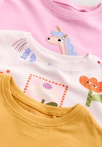 Trois chemises pour enfants à col rond : rose avec un écusson licorne, blanche avec des imprimés floraux colorés, et jaune moutarde uni.