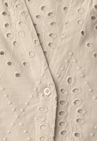 Tissu beige avec des motifs circulaires et ovales découpés brodés, avec un petit bouton blanc avec texte.