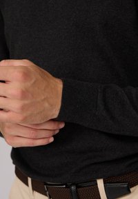 Pull en tricot gris foncé avec des poignets côtelés, présentant une coupe ajustée. Les mains tiennent la zone des poignets. Porté avec une ceinture tressée marron.