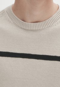 Helle beige Strickjacke mit einem gerippten Rundhalsausschnitt, die einen markanten horizontalen schwarzen Streifen über der Brust aufweist. Weiche Textur.
