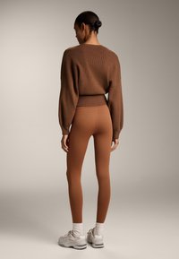 Pulover tricotat maro cu un tiv texturat, asortat cu colanți maro și pantofi sport albi. Întreaga ținută are un design texturat și o siluetă ajustată.