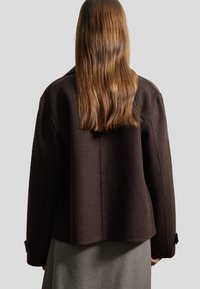 Bruine wolmix jas met een relaxte pasvorm, brede schouders en een iets verkorte lengte. Bevat knoopmanchetten en minimale stikseldetails.