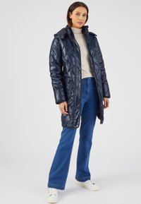 Cappotto puffer trapuntato blu navy con colletto alto, bottoni a pressione e tasche laterali. Indossato sopra un dolcevita beige chiaro e jeans di denim.