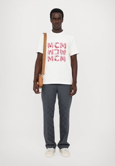 T-shirt en coton blanc avec un texte graphique rose, porté avec un pantalon gris à motifs et des baskets blanches. Sac à dos marron avec une bandoulière.
