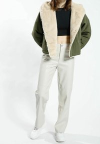 Olivgrön jacka med en faux fur-krage, kombinerad med ljusbeige byxor. Jackan har en kortad design och tryckknappar.