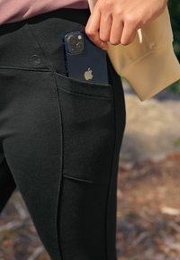Des leggings noirs en tissu lisse et extensible, dotés d'une poche latérale tenant un smartphone, et d'un petit logo près de la taille.