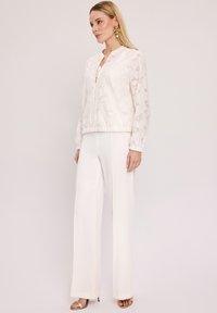 Biała kurtka bombowiec o teksturowanej tkaninie i zamek błyskawiczny, zestawiona z białymi spodniami o wysokim stanie, dobrze dopasowanymi oraz złotymi sandałami na obcasie.