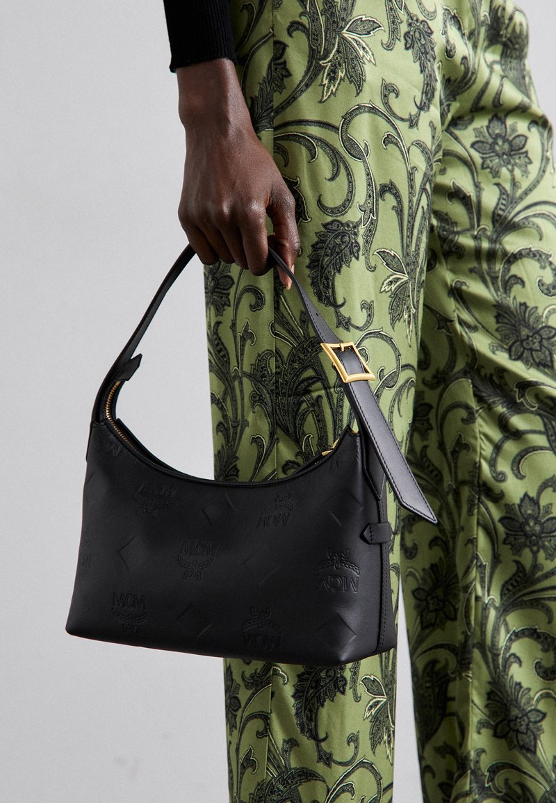 Mustat olkalaukku, jossa on kohokuvioitu logomalli, sileä nahkamateriaali, kaareva muoto ja kultainen kiinnitysremmissä.