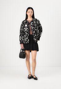 Casaco oversized em padrão preto e branco com decote redondo, usado sobre uma blusa justa castanha, combinado com uma saia preta com babados e uma pequena mala.