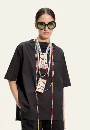 Kobieta w luźnej czarnej koszuli, nosząca duże okrągłe okulary przeciwsłoneczne i wiele długich koralikowych naszyjników z saszetkami w zwierzęcy wzór.