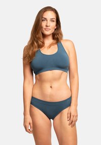 Turkusowy stanik sportowy i pasujące majtki bikini; gładki materiał, dopasowany krój, żebrowana faktura i szerokie ramiączka. Modelka stoi przodem.