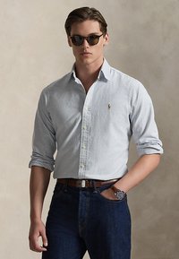Mann som har på seg en lyseblå stripete skjorte med knapper, mørke jeans, brunt belte, armbåndsur og solbriller mot en nøytral bakgrunn.