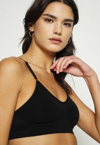 Μαύρο σουτιέν μπράλετ από μαλακό ύφασμα, με στρογγυλή λαιμόκοψη, ρυθμιζόμενα λουράκια και πτυχές στο κέντρο για άνετη εφαρμογή.