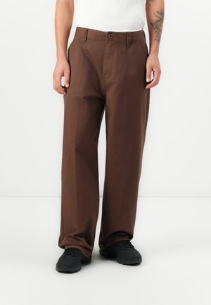 Brown, wide-legged trousers made of cotton with a smooth texture. Features a button closure and belt loops. Worn with black sneakers.