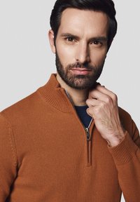 Brązowy sweter zapinany na zamek z wysokim kołnierzem, o teksturowanym wzorze dzianiny, z prążkowanymi mankietami. Nosi go na granatowej koszuli. Model dotyka kołnierza.