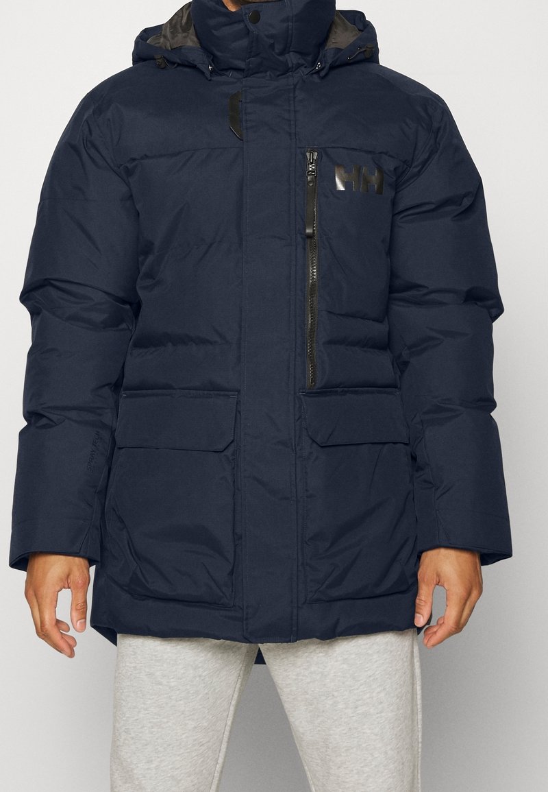 Mørkeblå isoleret parka med lynlås foran, store lommer og hætte. Har et sort logo på brystet og struktureret stof.