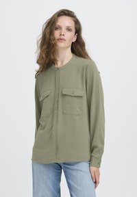 Jeune femme aux longs cheveux ondulés portant une chemise légère olive à boutons avec des poches poitrine et un jean bleu clair, se tenant devant un fond blanc.