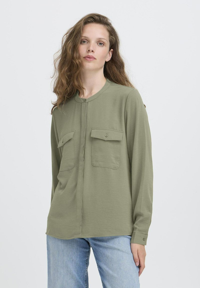 Jeune femme aux longs cheveux ondulés portant une chemise légère olive à boutons avec des poches poitrine et un jean bleu clair, se tenant devant un fond blanc.