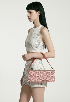 Jonge vrouw met een bloemenprint cropped top en shorts houdt een roze handtas met patroon en rode band vast tegen een effen achtergrond.