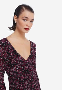 Schwarzes und burgunderfarbenes gemustertes Kleid mit tiefem V-Ausschnitt, langen Ärmeln und gerafften Details. Das Model trägt silberne Herzohrringe.