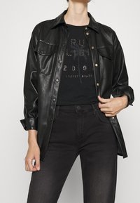 Chaqueta de camisa de cuero negro con botones a presión, dos bolsillos en el pecho y puños enrollados, llevada sobre una camiseta negra con un brillante estampado gráfico.