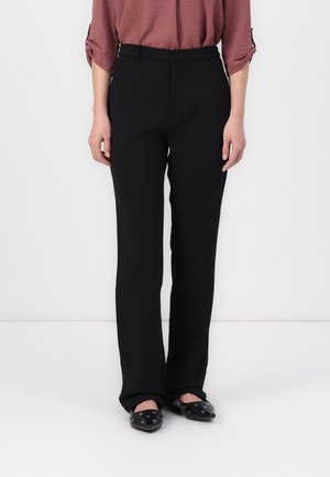 Personne portant un pantalon noir taille haute à jambes droites, associé à des ballerines noires pointues et un haut à manches longues mauve, debout à l'intérieur.