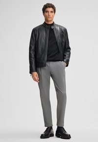 Giubbotto di pelle nera con spalle rigate, dolcevita nero, pantaloni grigi a fantasia e scarpe nere stringate. Design semplice e su misura.