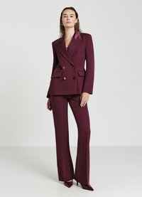 Donna in piedi che indossa un blazer bordeaux su misura a doppio petto e pantaloni larghi coordinati, con tacchi a punta.