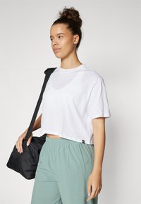 T-shirt bianca cropped con scollo rotondo, abbinata a pantaloni verde chiaro. La modella tiene una borsa da viaggio nera con tracolla.